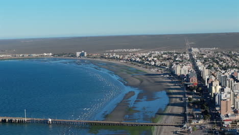4k 60fps drone shot over puerto madryn’s coastline. dock and building shadows cast on the shoreline under a clear blue sky. distant horizon.