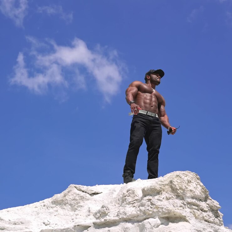 Torso desnudo de atleta afroamericano. Culturista afroamericano en la cima de una colina blanca con un fondo de cielo azul. Vista desde abajo.