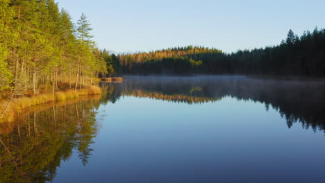 Drone-flying-low-above-a-calm-misty-lake-by-beautiful-colorful-autumn-morning