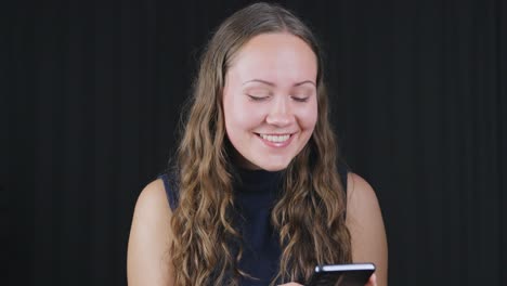 Mujer-Atractiva-En-Sonrisas-Enviando-Mensajes-De-Texto-Tocando-El-Teléfono-Contra-El-Fondo-Negro