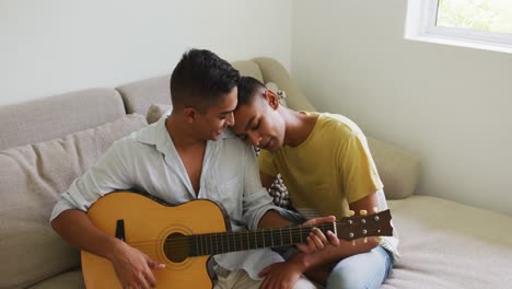 smiling mixed race gay male couple sitting on sofa and embracing while one plays guitar