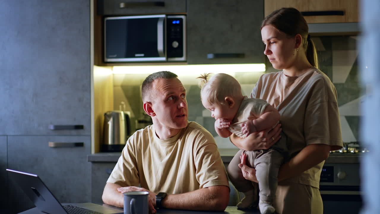 Woman with a child in hands stands behind her husband sitting at the table. Man looks at laptop and says something to his wife.
