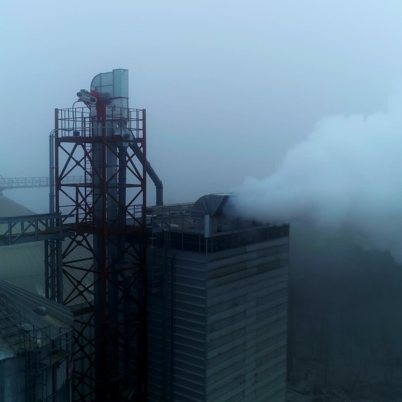 Approaching the high building at elevator plant emitting thick white smoke. Aerial view. Dirty tops of granary tanks at agribusiness enterprise. Foggy day backdrop