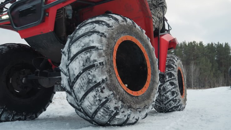 Primo piano di un pneumatico atv coperto di neve con pilota caucasico che si prepara per una corsa estrema, dettaglio del cerchio arancione, battistrada profondo intasato di brina, inquadratura dal basso che enfatizza la trazione e la forza meccanica.