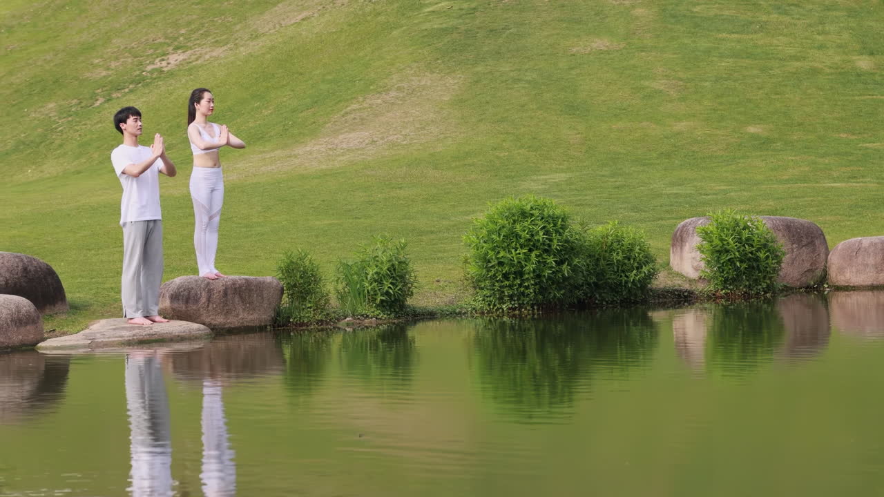 Giovane coppia spirituale e calma in piedi vicino al lago di un parco verde con gli occhi chiusi in posizione del loto, che pratica la meditazione respiratoria yoga, eseguendo il gesto namaste e tenendo le mani in mudra mentre si allena con esercizi yoga