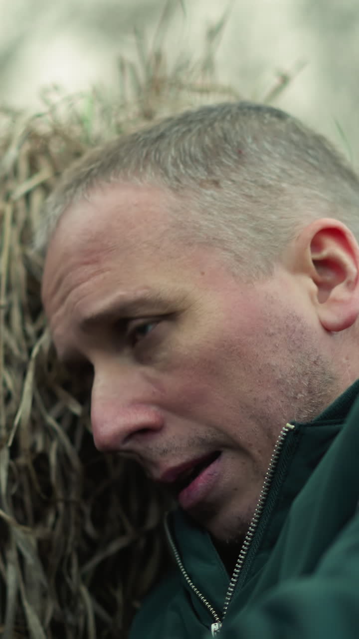 близкий взгляд на человека в зеленой куртке, лежащего в канаве против сухой травы с пистолетом в руке, человек выглядит измученным и напряженным