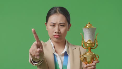 close up of asian business woman in a suit with a gold medal and trophy disapproving with no index finger sign and smiling to camera on green screen background in the studio