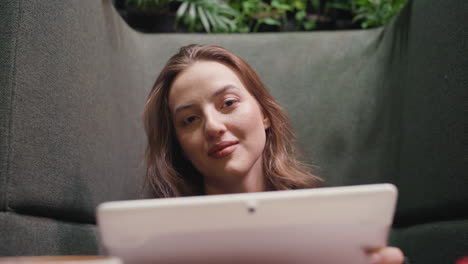 smiling lady holds notepad and looks directly at camera while seated on cushioned booth, surrounded by soft fabric panels and greenery, exuding peaceful energy and quiet self-assurance