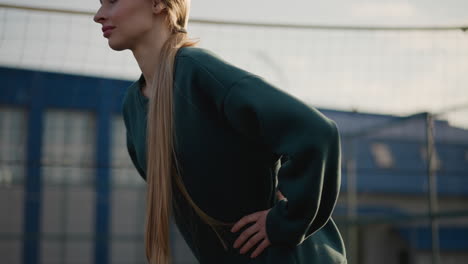 dama con sudadera verde y cabello largo realizando giros de cintura en la cancha al aire libre con el fondo borroso del edificio residencial y la red, capturando el estilo de vida saludable y la rutina de fitness