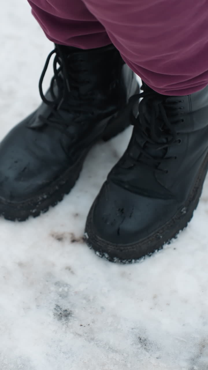 perspectiva de arriba hacia abajo de un individuo con botas negras saltando juguetón en el suelo cubierto de nieve con follaje seco esparcido, el entorno de invierno destaca el movimiento, la textura del calzado y la superficie helada