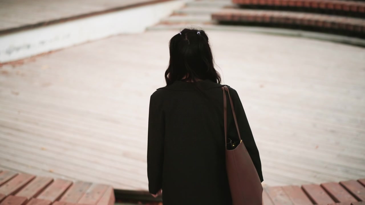 Young woman with long dark hair walks away from camera on wooden deck carrying brown bag over her shoulder. Woman wears black coat and white pants as woman moves through outdoor public space