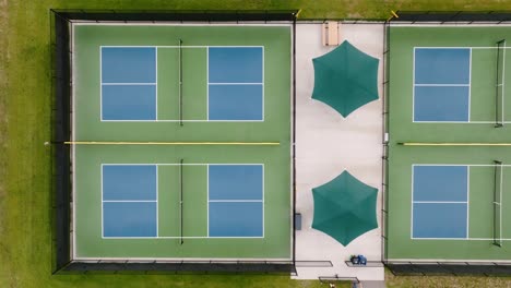 a top down aerial drone shot of four pickleball courts in a park with a nice seating area covered by two green umbrellas