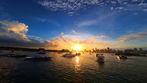 Multiple yachts stand on the anchor in the bay at sunset. FPV drone flies among the boats. Skyline of a big metropolis at backdrop.