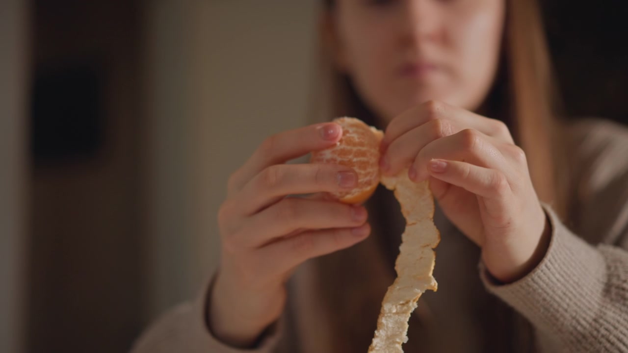 Caucasian woman peeling mandarin with focus closeup on hands removing rind delicate segments exposed juice glistening neutral kitchen background evening snack ritual mindful eating tactile detail slow