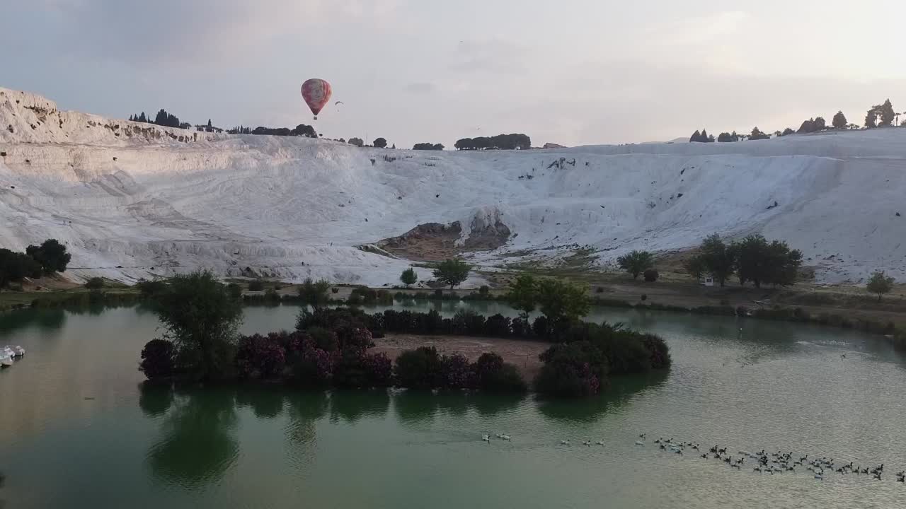 Türkiye, Pamukkale'de, traverten oluşumlarıyla çevrili gölün havadan görünümü, arka planda uçan sıcak hava balonuyla.