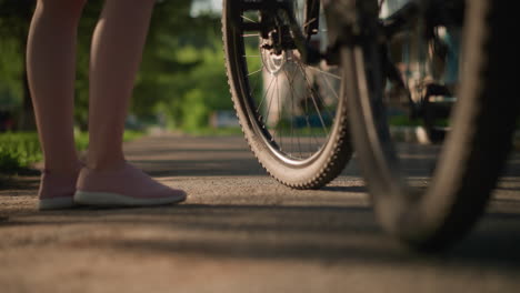 primer plano de piernas individuales en zapatillas rosadas pateando un neumático de bicicleta para comprobar la presión del aire, la vegetación exuberante y los árboles crean un hermoso telón de fondo, mientras un coche pasa en el fondo lejano