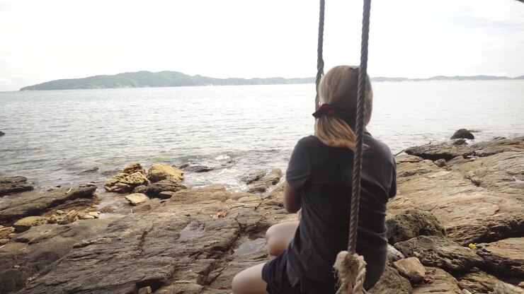 Una viaggiatrice siede su un'altalena di legno rustica con vista sulla costa rocciosa e sul vasto oceano al Parco Nazionale Khao Laem Ya Mu Ko Samet, ammirando le serene vedute dell'isola.