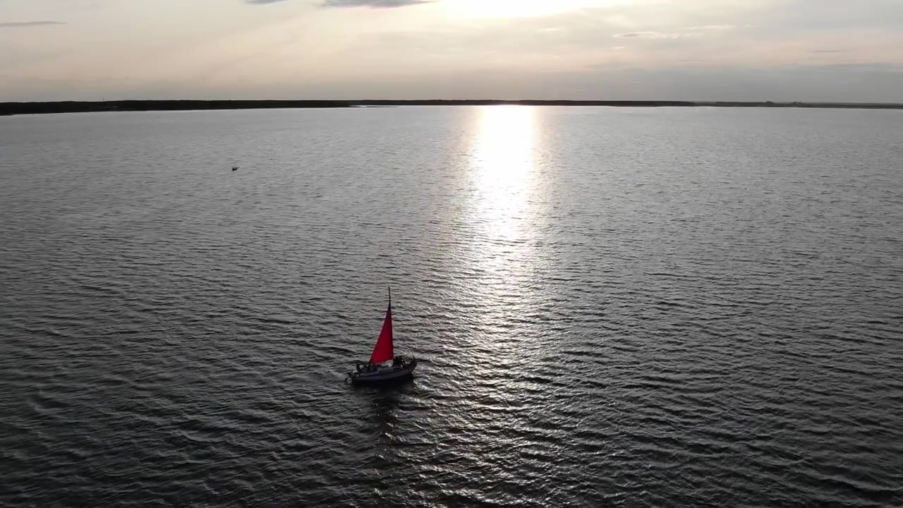 Vista aérea de velero en río al atardecer, siguiendo hacia el horizonte.