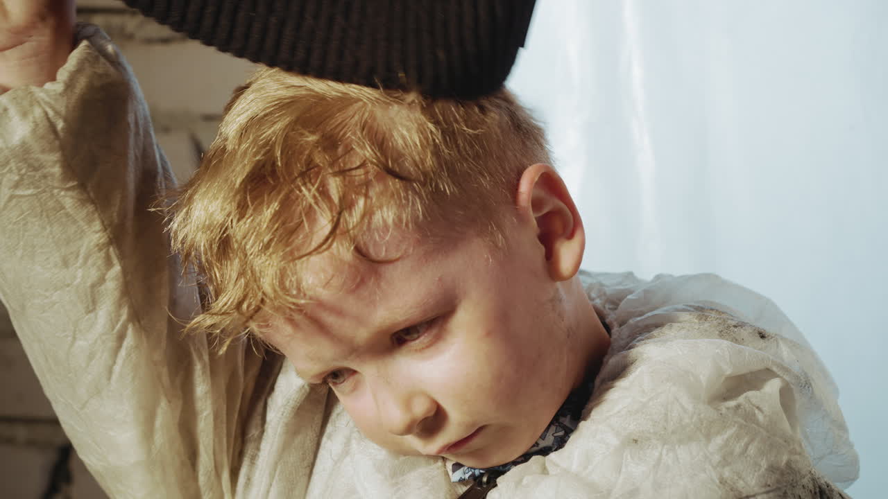 Close up of young boy in dirty protective suit pulling black beanie over head while sitting near cracked brick wall and plastic tarp, face tired and dusty