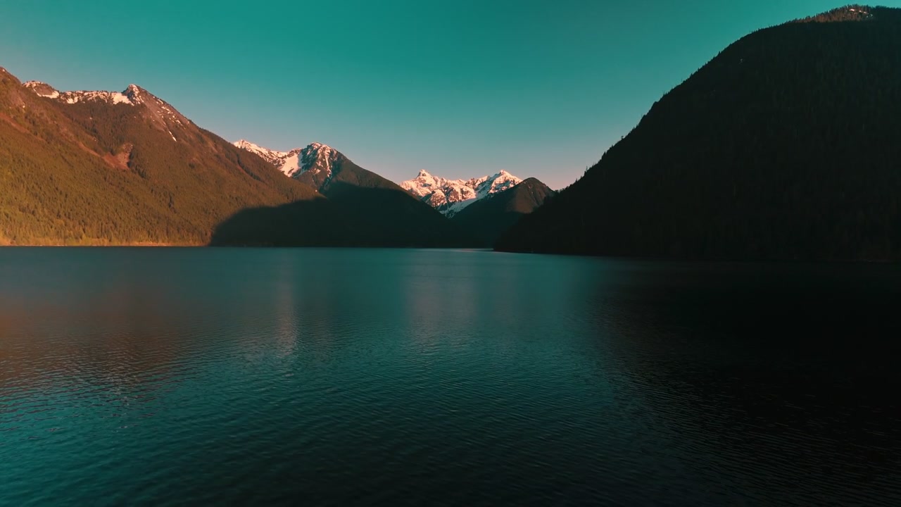 Ampia vista attraverso l'acqua scura, calma e increspata del lago di fronte a una maestosa catena montuosa aspra con cime innevate che brillano intensamente con la luce dorata e brillante del tramonto, ripresa aerea con drone in avvicinamento ascendente