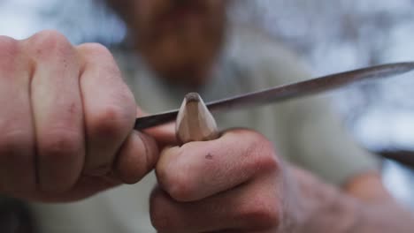 caucasian male survivalist cutting point in stick to make fire drill at camp in wilderness