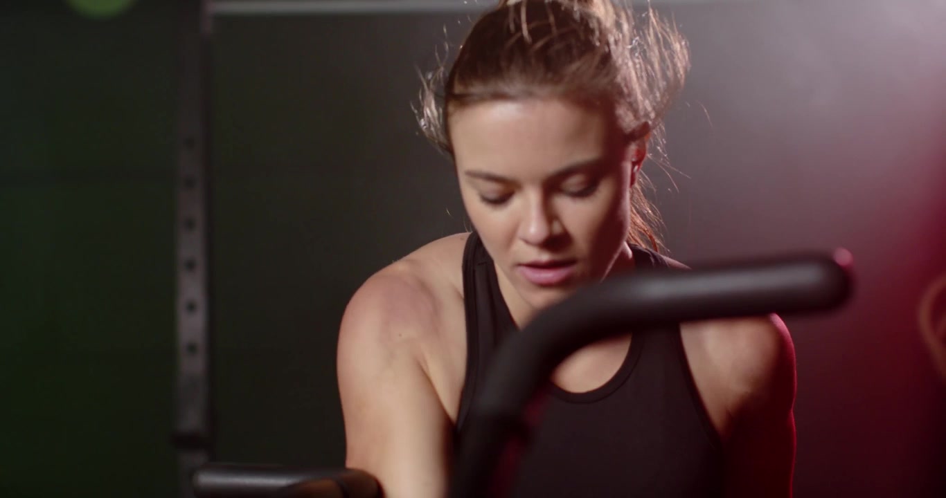Woman starting interval, pedaling bike in gym, leaning on bars, eyeing console in black tank top