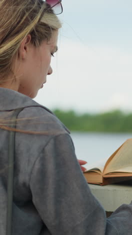 dama de camisa gris leyendo un libro mientras descansa en una valla, el viento sopla suavemente las páginas mientras permanece concentrada, con un fondo de agua tranquila y vegetación