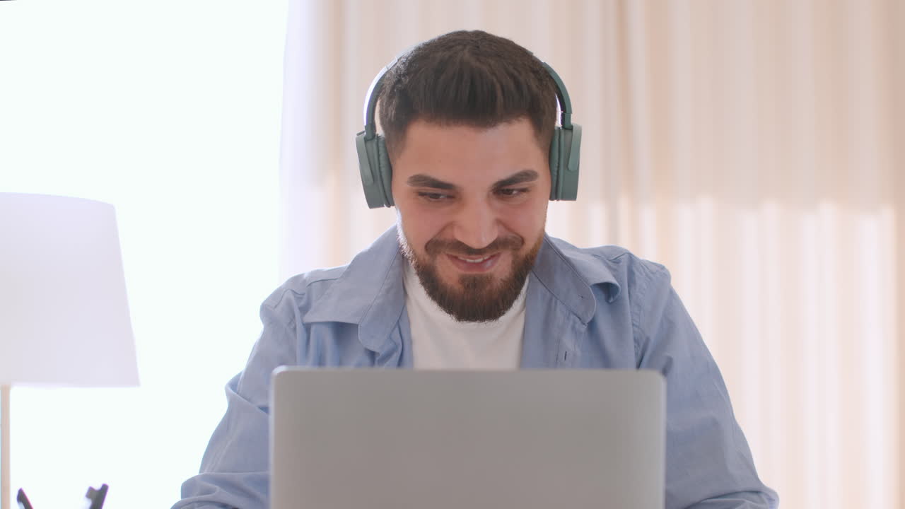 Bearded man wearing headphones uses a laptop at home