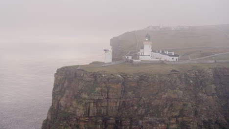 Luftaufnahme-Eines-Nebligen-Leuchtturms,-Der-In-Der-Abenddämmerung-Auf-Einer-Klippe-Am-Meer-Leuchtet