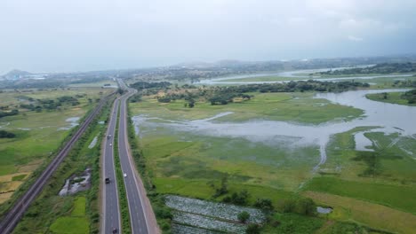 cinematic aerial drone shot of vast farming land flooded in water beside passing cars and heavy vehicles on the four lanes highway and a railway track