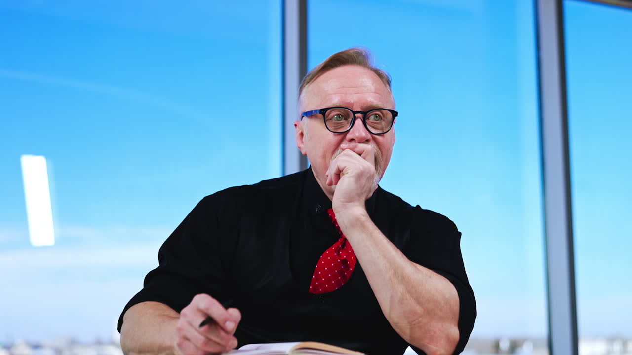 Adult Caucasian man in black clothes and red tie sits at desk. Businessman stops writing and sits still thinking.