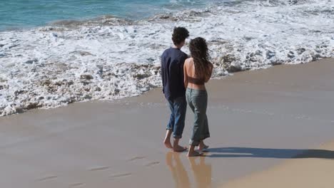romantic couple holding hands while walking barefoot on sandy shoreline. aerial tracking shot