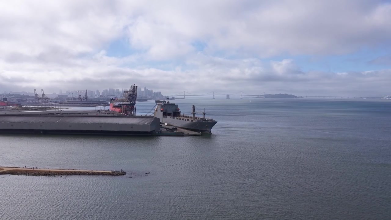 Cinematic drone video captures expansive Heron's Head Park waterfront, industrial piers, ships, calm bay waters and the distant San Francisco skyline under scattered clouds in soft daylight.