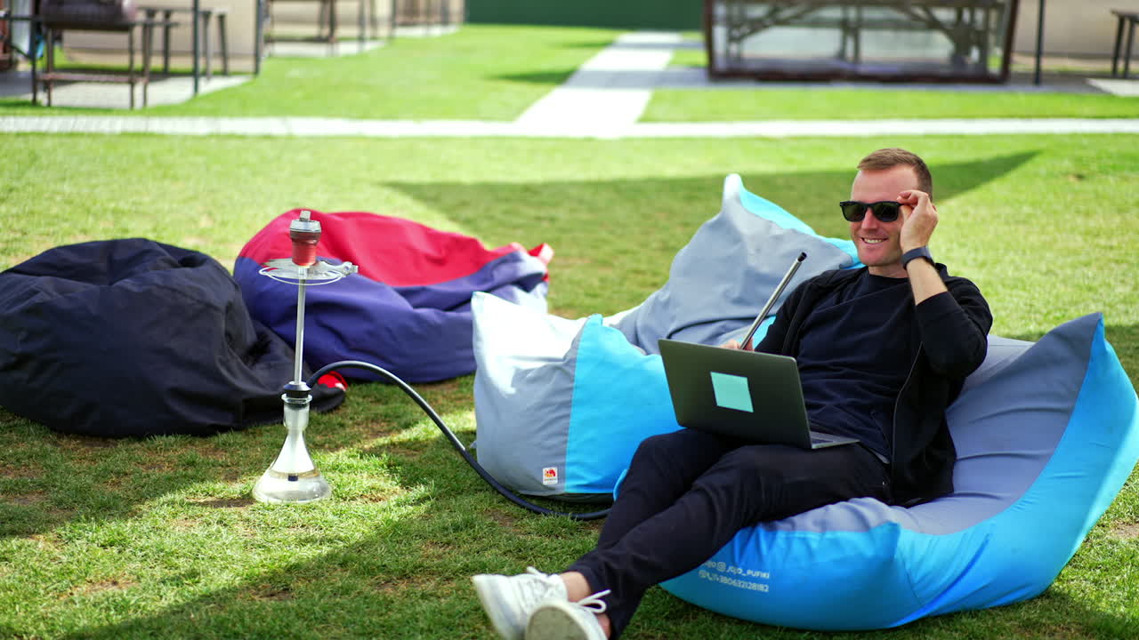 Smiling man in chair outdoors puts on sunglasses. Male finishing work on laptop and stars smoking hookah leaning in the chair.