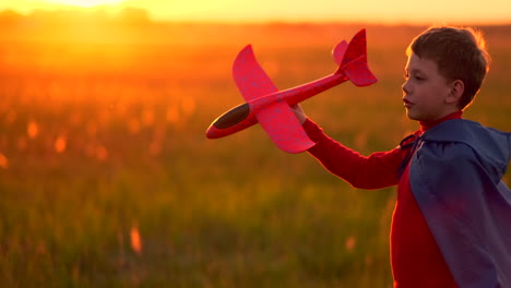 Der-Junge-Im-Kostüm-Eines-Superhelden-Läuft-In-Einem-Roten-Mantel-Und-Lacht-Bei-Sonnenuntergang-Auf-Einer-Sommerwiese-Und-Stellt-Sich-Vor,-Er-Sei-Der-Pilot-Des-Flugzeugs-Und-Spiele-Mit-Einem-Modellflugzeug.