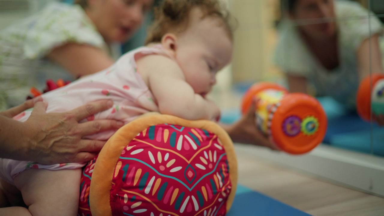 Bebê fazendo tempo de bruços em um tapete de atividades com um rolo de terapia, recebendo assistência suave para o desenvolvimento de habilidades motoras.