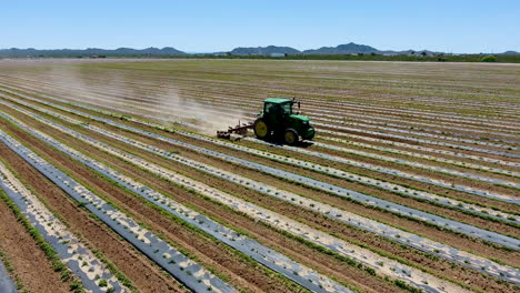 Wide-rotating-drone-shot-of-tractor-in-a-field-with-rows-of-crops,-with-mountains-in-the-background