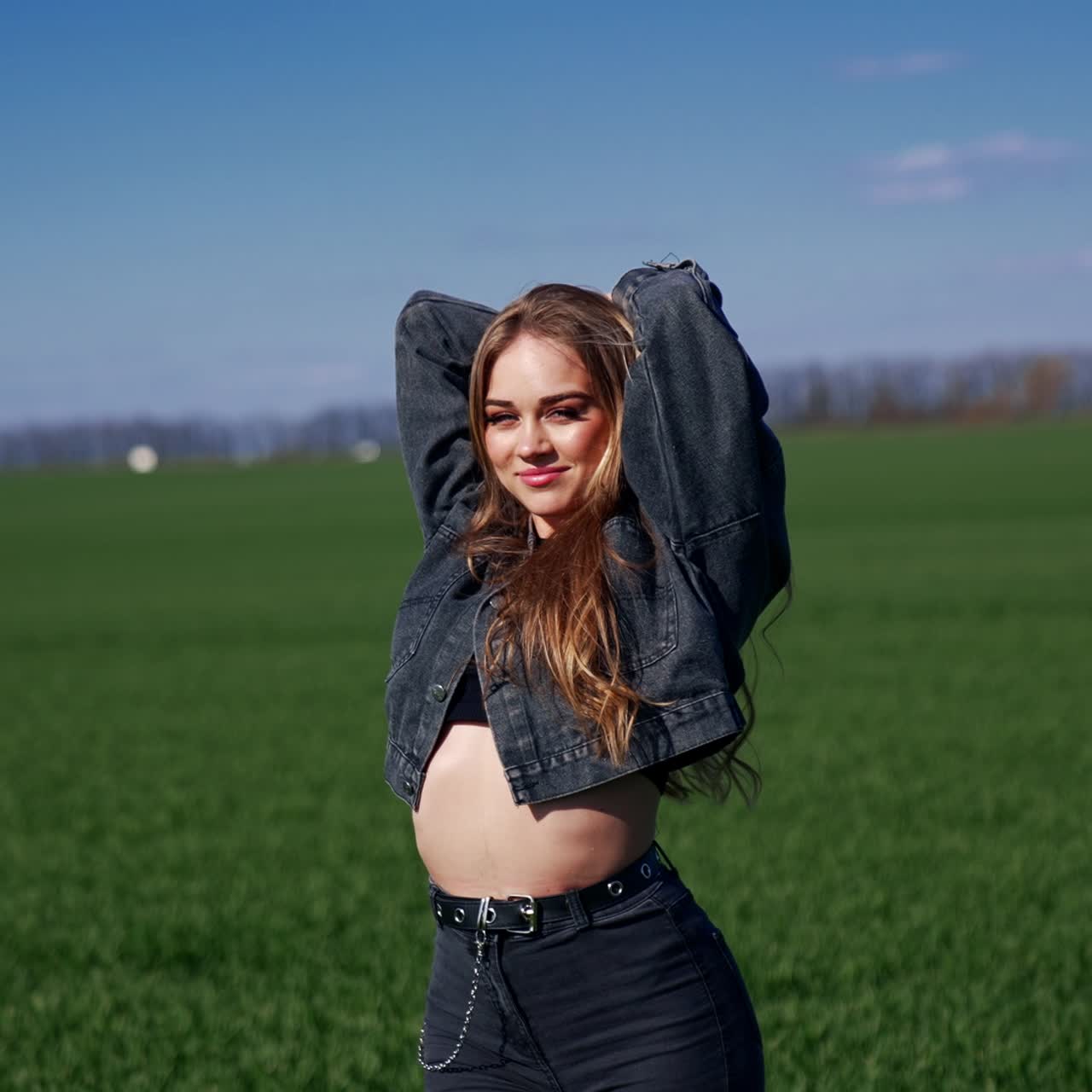 Sexy model posing on camera outdoors. Portrait of a beautiful young woman with long hair in short top and denim suit on green field in sunny day. Slow motion