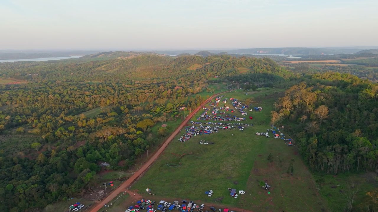 Ripresa aerea con drone ad alta quota di un vasto raduno di campeggiatori durante l'ora d'oro a Nueva Alborada, Paraguay. Centinaia di tende punteggiano un ampio paesaggio verde.