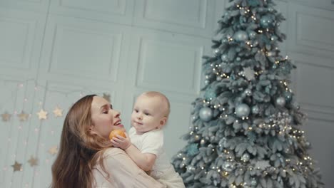 Cheerful-mother-kissing-cute-child-near-christmas-tree-in-modern-apartment.