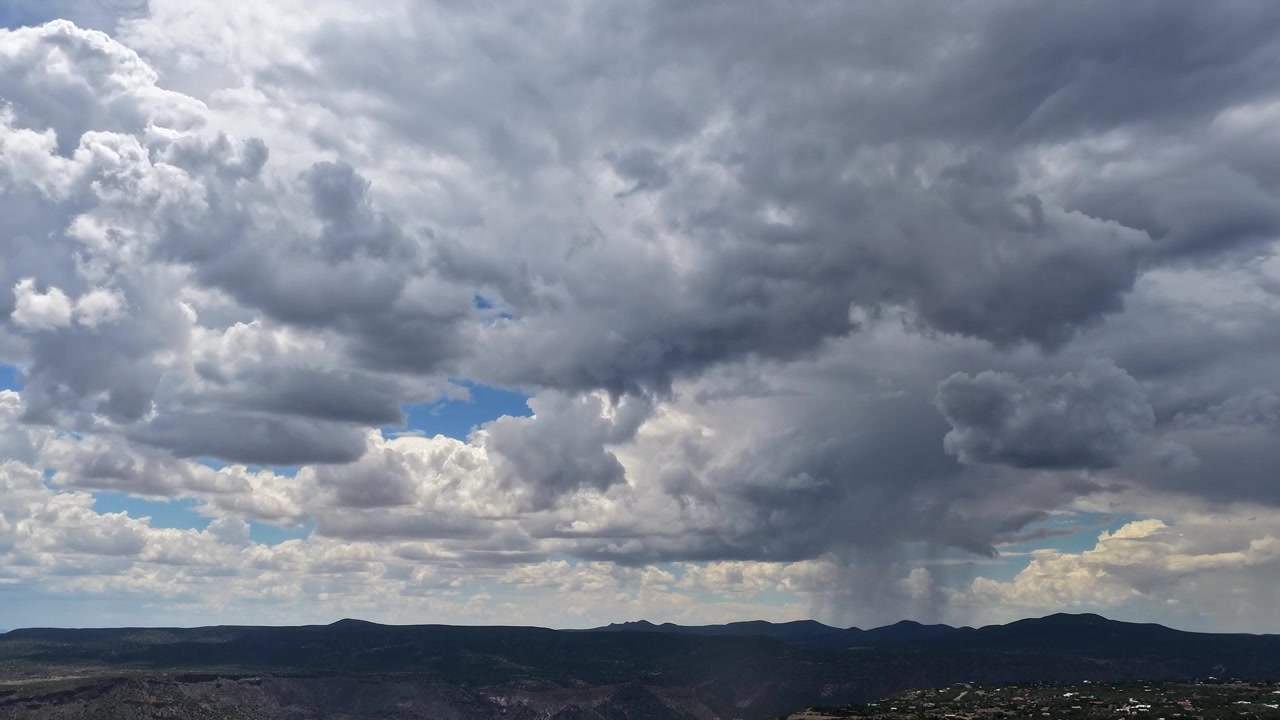 Timelapse 4K con drone che osserva un temporale localizzato su un canyon desertico elevato. Squarci di cielo blu si fanno strada tra nuvole temporalesche scure e pesanti mentre una colonna di pioggia visibile inzuppa il lontano terreno accidentato