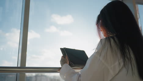 side view of lady operating tablet near large glass window with cityscape and sunlight illuminating her hair and hands, creating warm modern tech inspired scene with subtle reflections and shadows