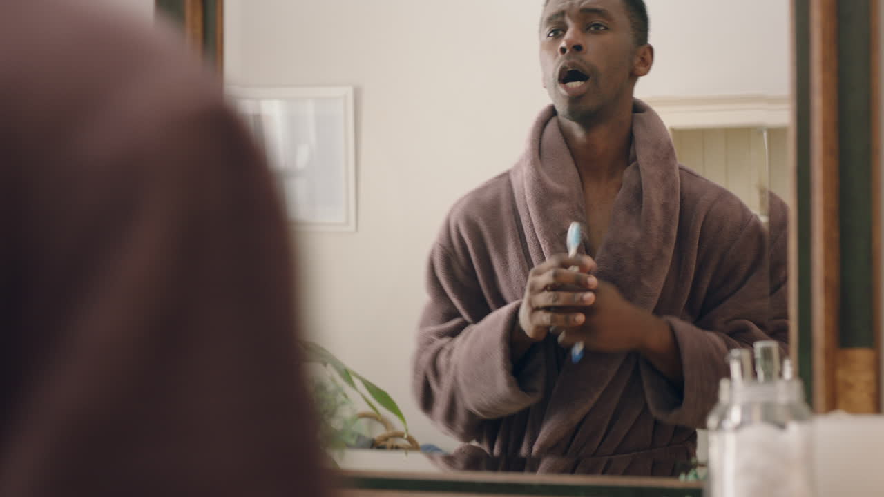 행복한 아프리카계 미국인 남자가 화장실에서 춤을 추고 거울을 보며 즐거운 아침 루틴을 준비하고 긍정적인 자기 이미지를 즐기고 목욕옷을 입고 있습니다.