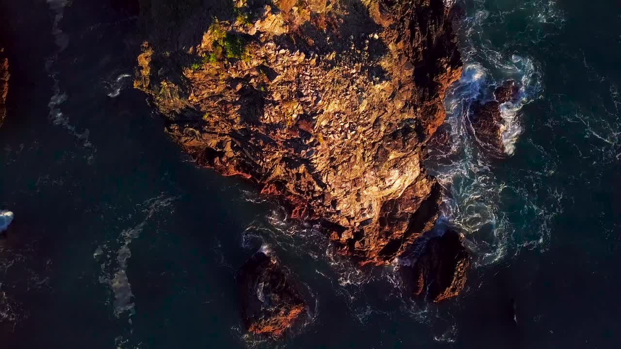 Vista aerea ascendente di scogliere rocciose e onde oceaniche spumeggianti al tramonto a Big Sur, California.