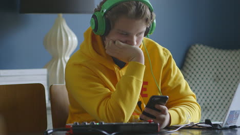 Teen-boy-on-iPhone-with-midi-controller-and-laptop-and-wearing-headphones,-at-home,-medium-shot