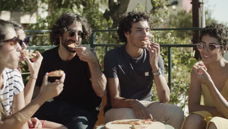 gente feliz sentada y comiendo pizza durante la fiesta en la azotea en un día ventoso de verano