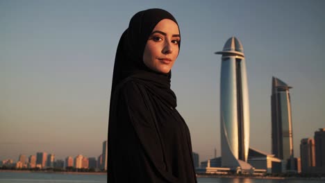 elegant businesswoman wearing a traditional black abaya and hijab is posing with jeddah's modern skyline and the prominent headquarters of a saudi arabian bank in the background during sunset