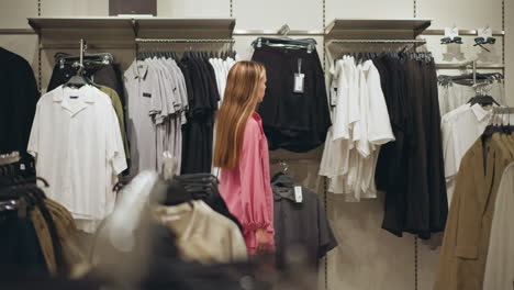 dama vestida con tela rosa camina pensativa a través de una tienda de ropa, deteniéndose para mirar una chaqueta marrón entre estantes bien dispuestos de camisas y chaquetas, tienda bien organizada con estantes y estantes