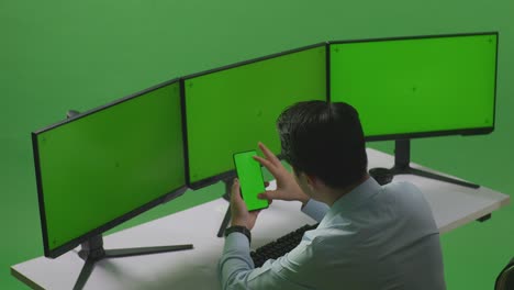 vista lateral de un hombre asiático usando un teléfono móvil y varios monitores de computadora con pantalla verde simulada en el estudio de pantalla verde