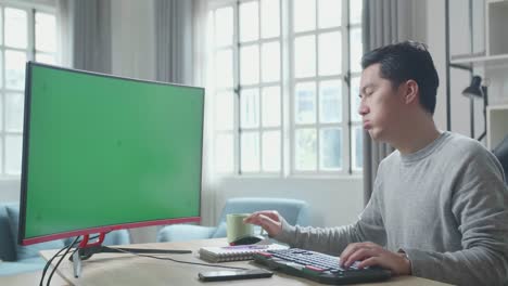 asian man in long sleeved t-shirt being tired while using green screen desktop computer for working at home.